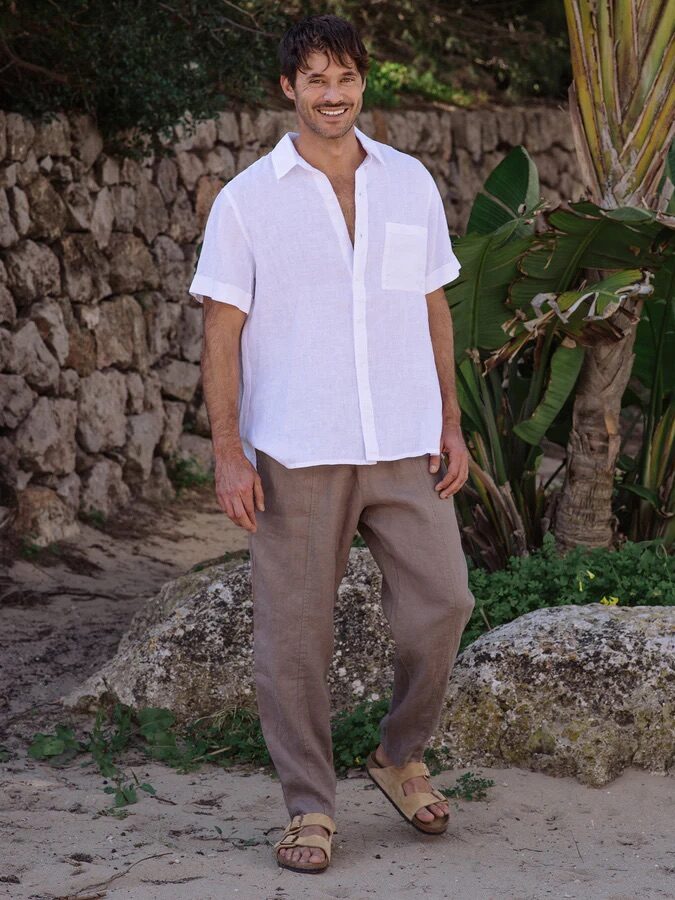 A man stands outdoors on a sandy path, wearing a white short-sleeve shirt, beige pants, and sandals, with greenery and a stone wall in the background.