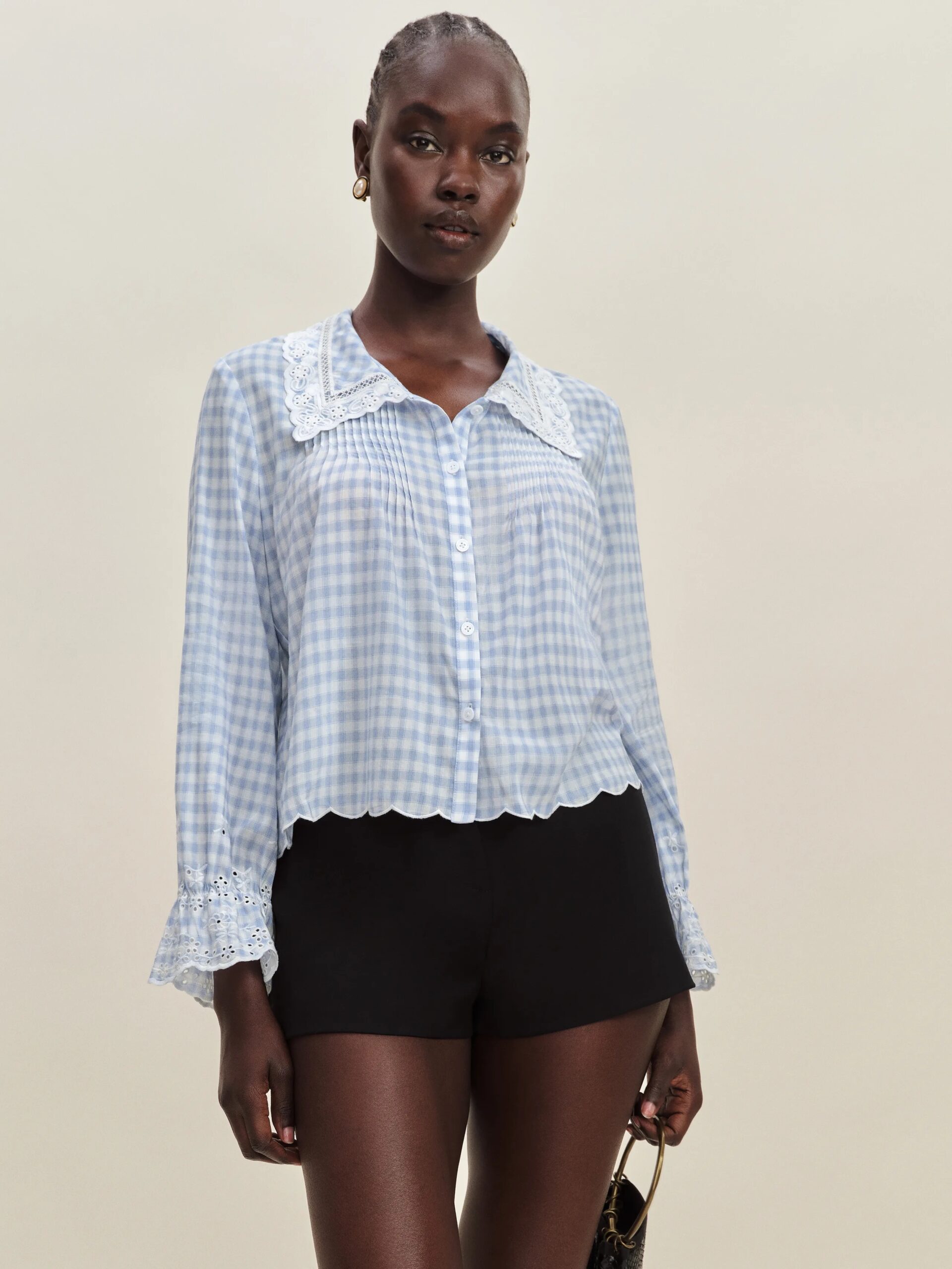 A woman poses against a plain background wearing a light blue gingham blouse with lace collar and cuffs, and black high-waisted shorts.