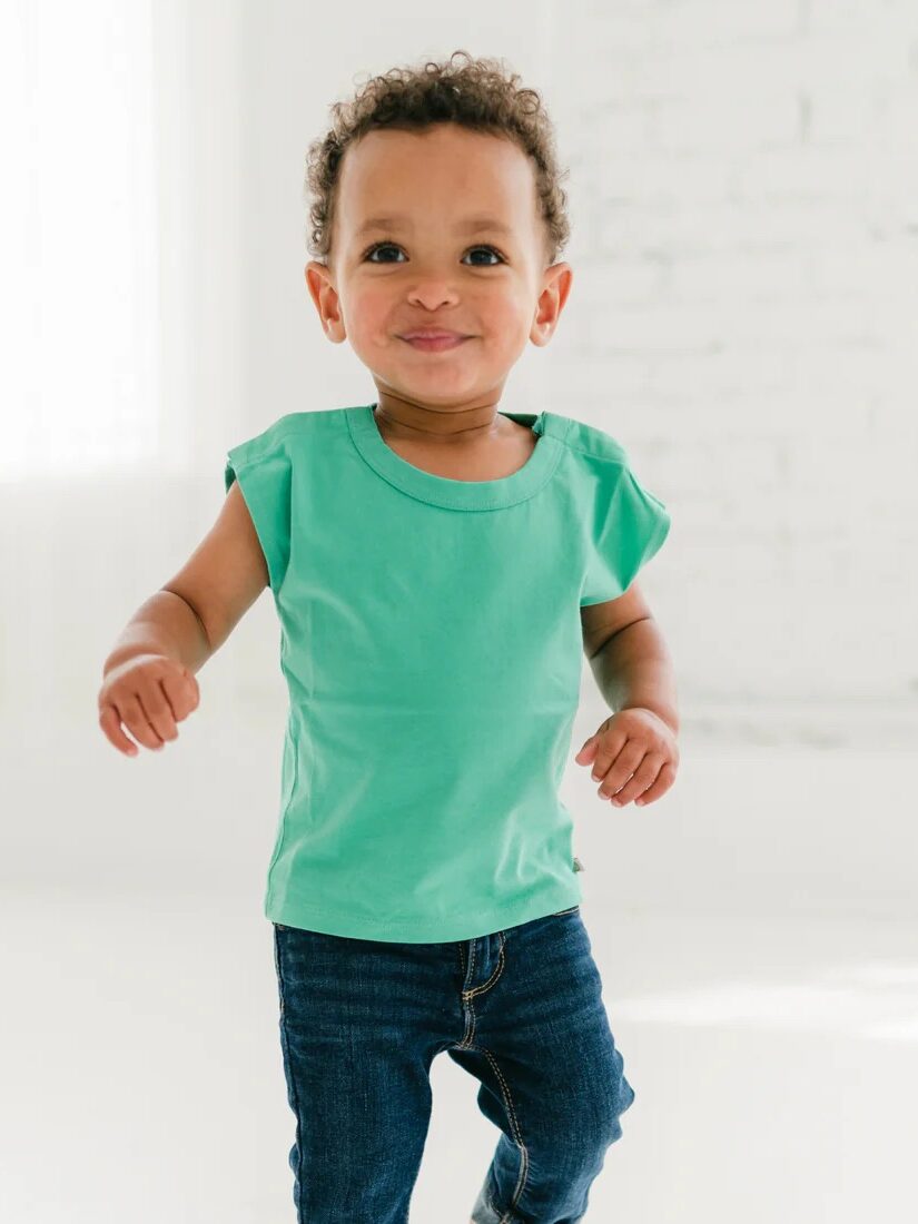 A toddler in a green t-shirt and blue jeans stands indoors on a white floor with a white brick wall in the background.
