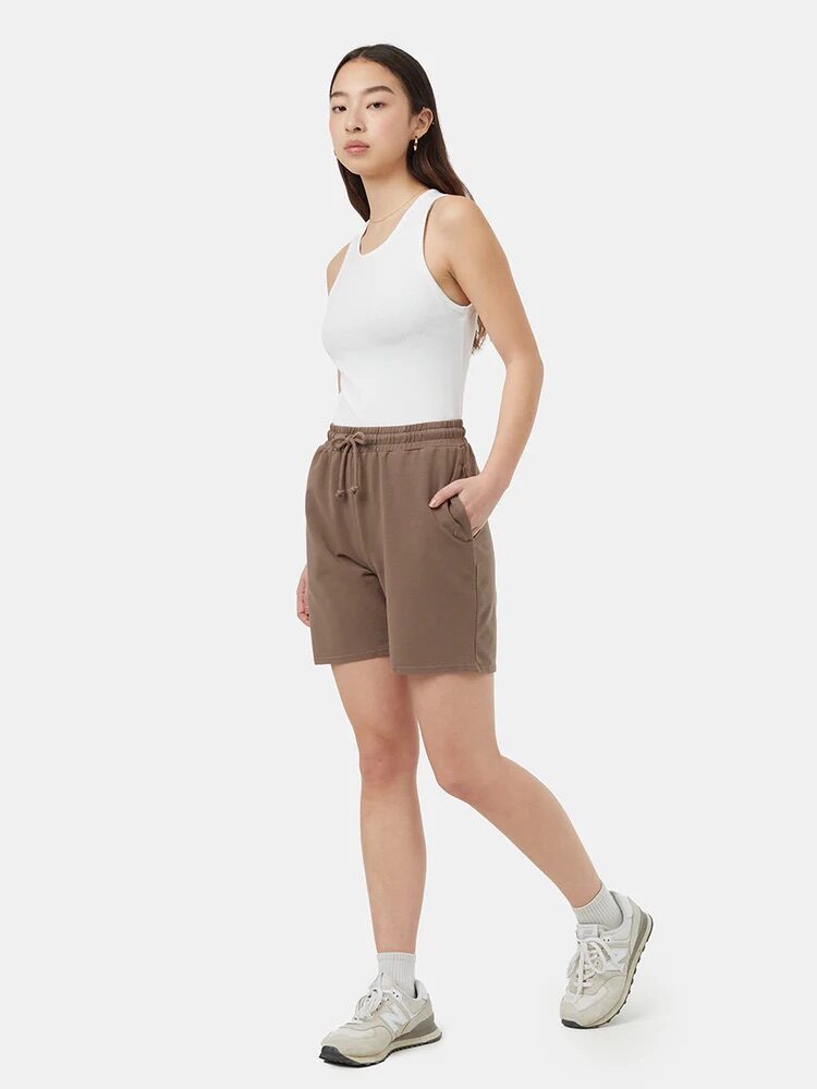 A woman stands against a white background wearing a white sleeveless top, brown drawstring shorts, white socks, and white sneakers.