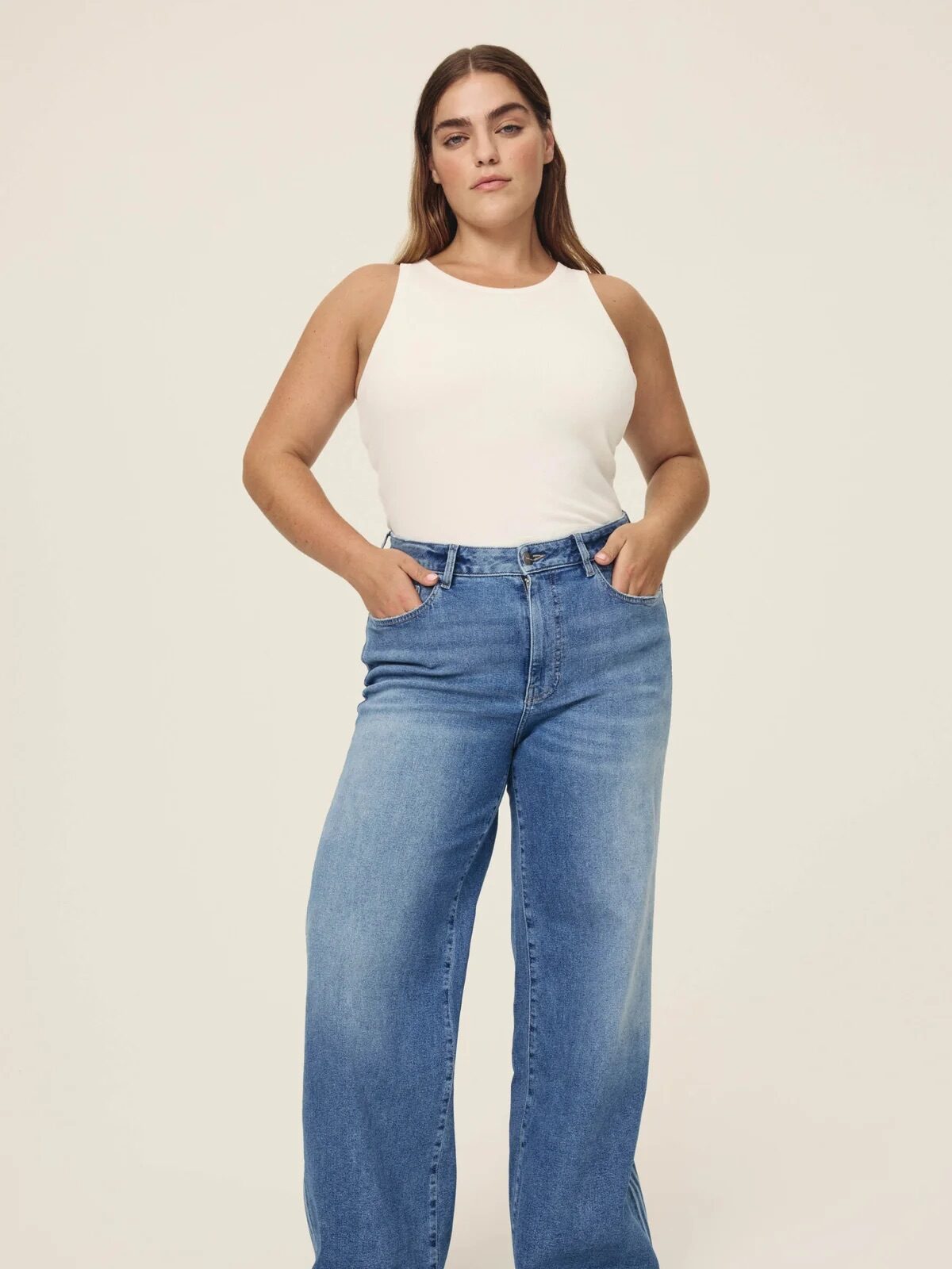 A woman stands facing forward, wearing a white sleeveless top and high-waisted wide-leg blue jeans with hands in her pockets, against a plain background.