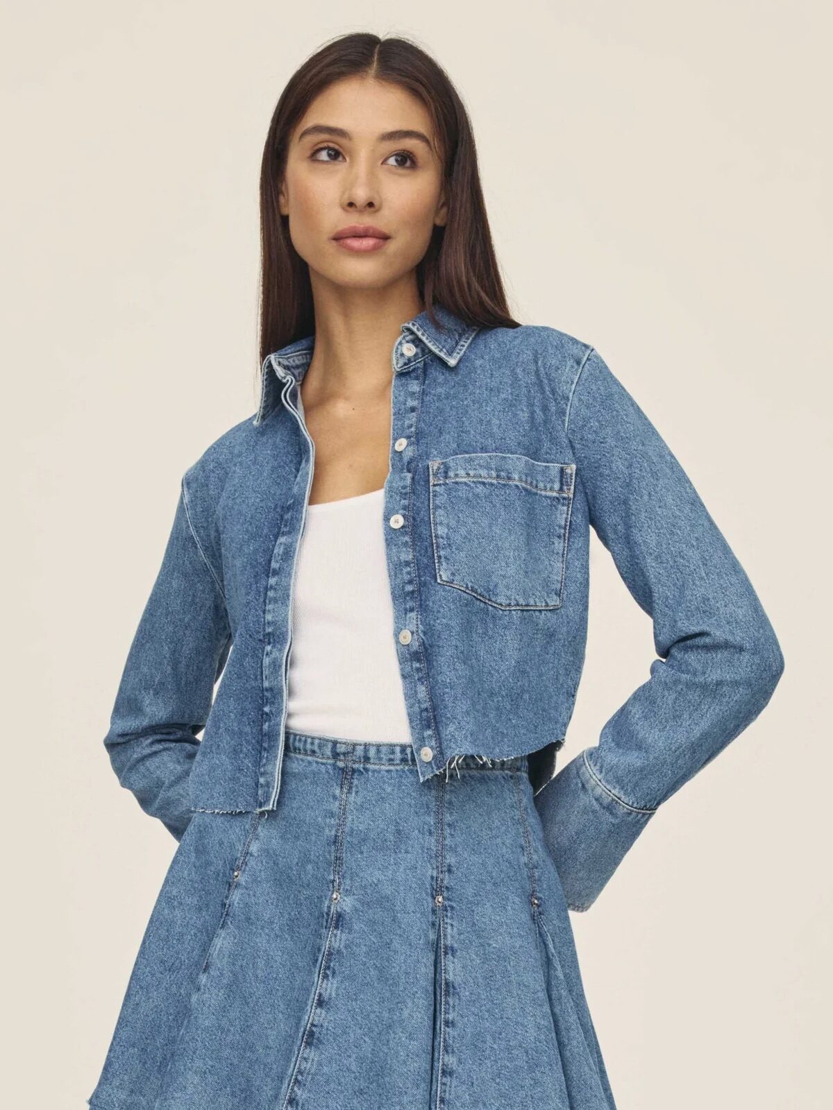 A woman wearing a cropped denim button-up shirt over a white top and a pleated denim mini skirt stands against a plain background.