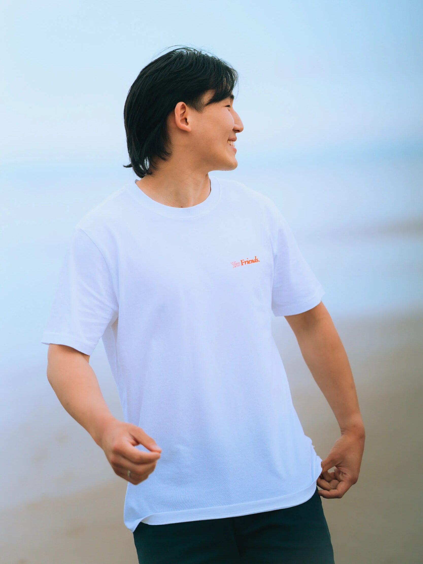 A person with medium-length dark hair wearing a white T-shirt stands outdoors, looking to the side with a slight smile. The background appears to be a beach or shoreline.