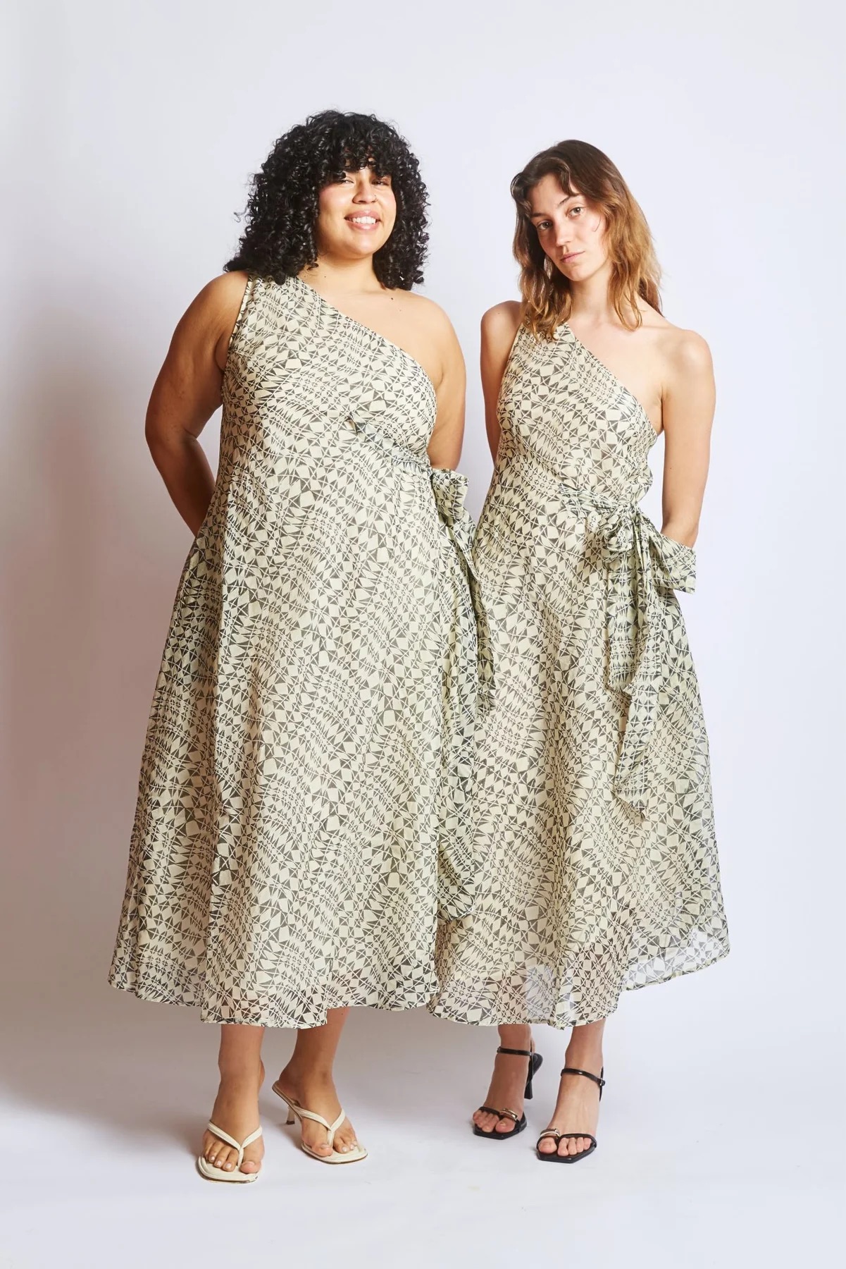 Two women wearing matching one-shoulder, ankle-length dresses with a geometric print and side bow, standing side by side against a plain background.