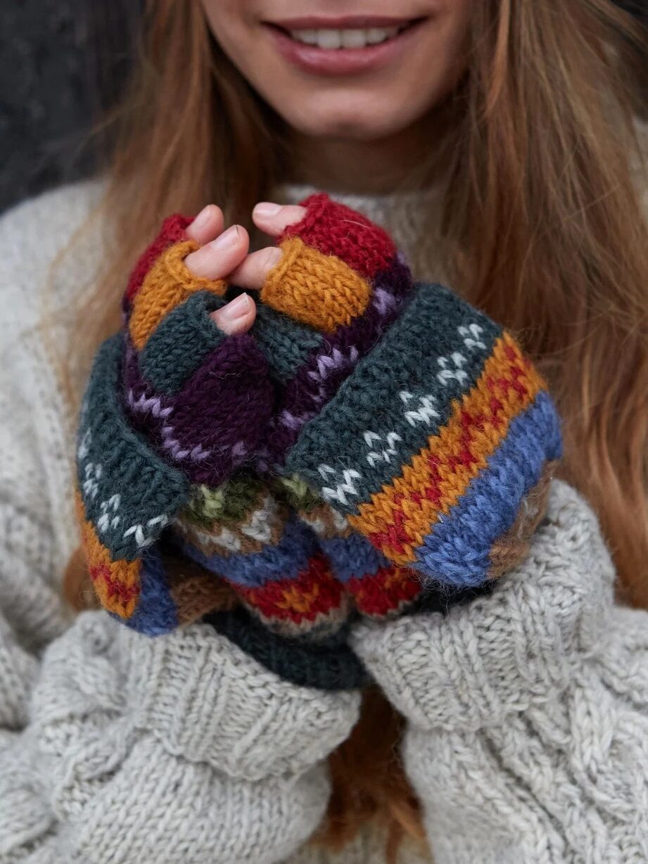 A person wearing colorful, patterned mittens and a chunky gray sweater holds their hands together, smiling softly.