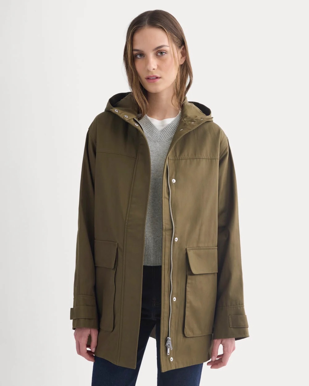 A woman wearing a waterproof olive green hooded raincoat over a gray sweater and dark pants stands against a plain white background.