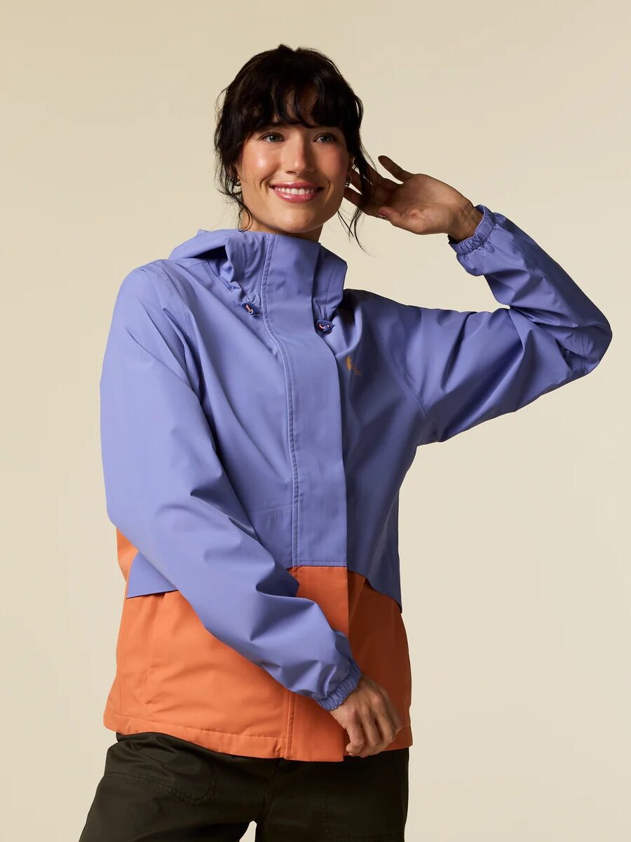 A person wearing a light purple and orange waterproof raincoat with olive green pants stands against a plain beige background, smiling and touching their hair.