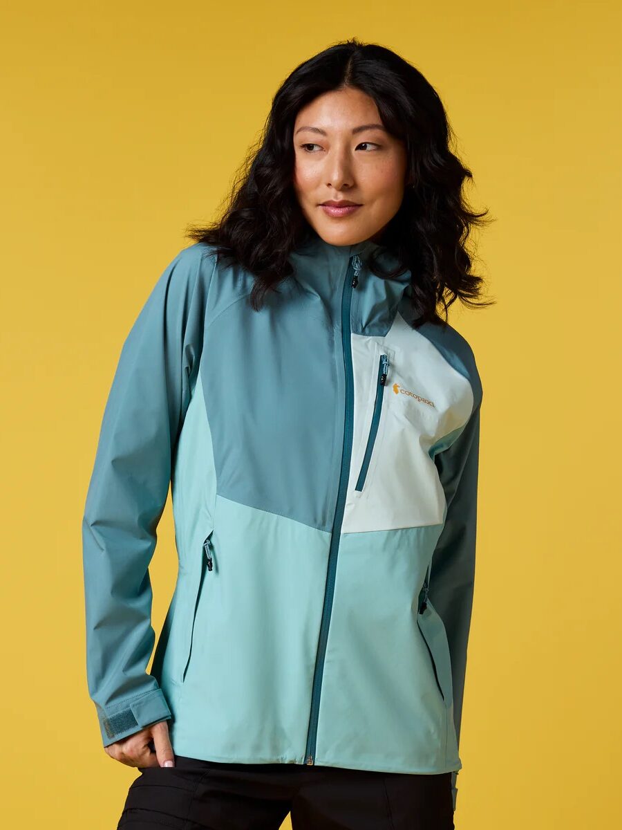 Woman wearing a teal and light blue waterproof raincoat stands against a plain yellow background, looking to the side with one hand in her pocket.