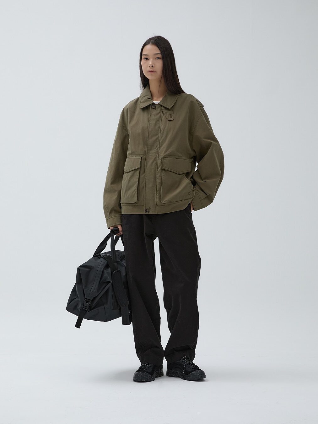 A person stands in a studio wearing a waterproof raincoat styled as an olive green jacket, black pants, and black shoes, holding a black duffel bag in their left hand against a plain white background.