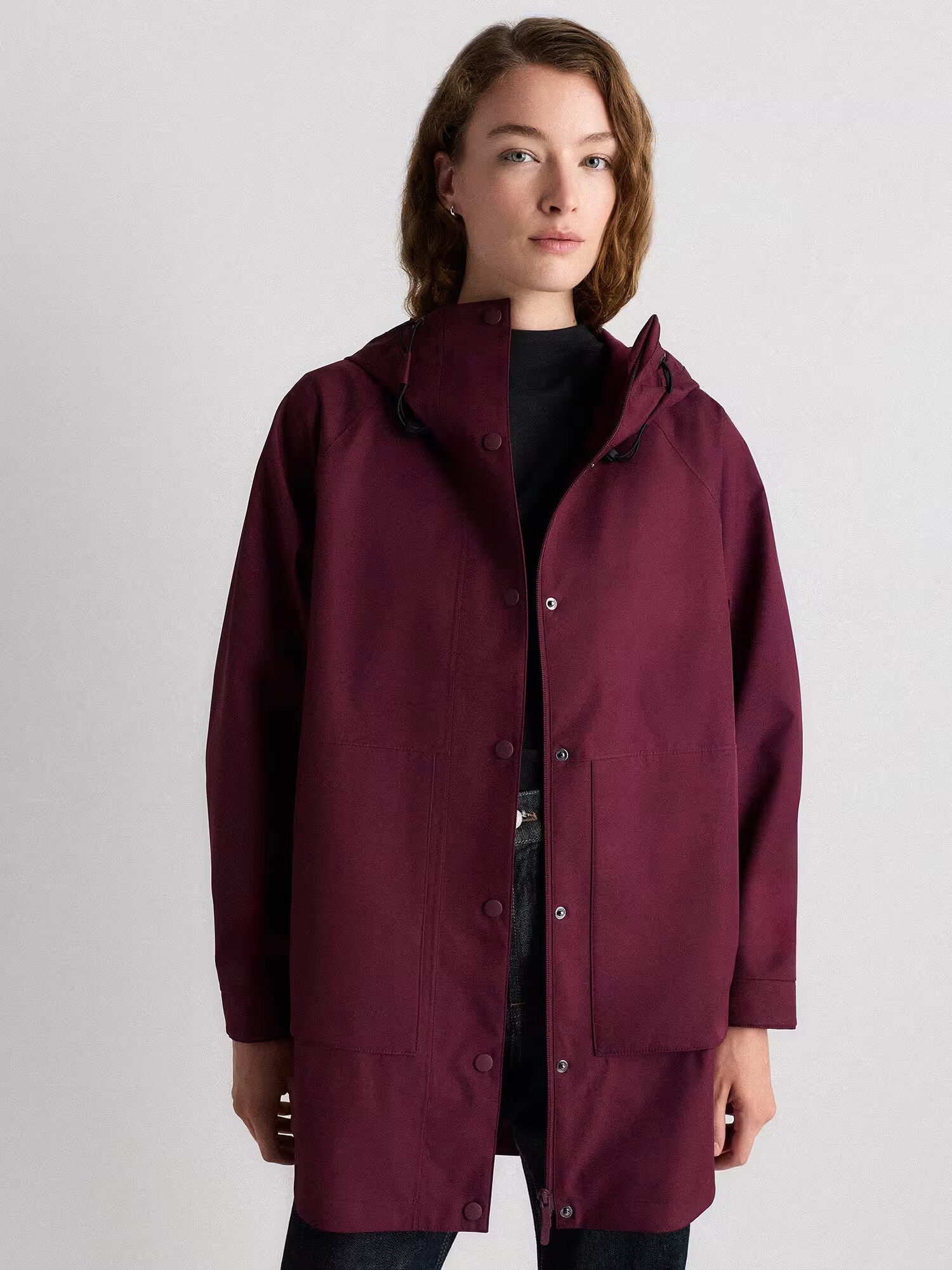 Person wearing a long, dark maroon waterproof raincoat over a black top, standing against a plain light grey background.