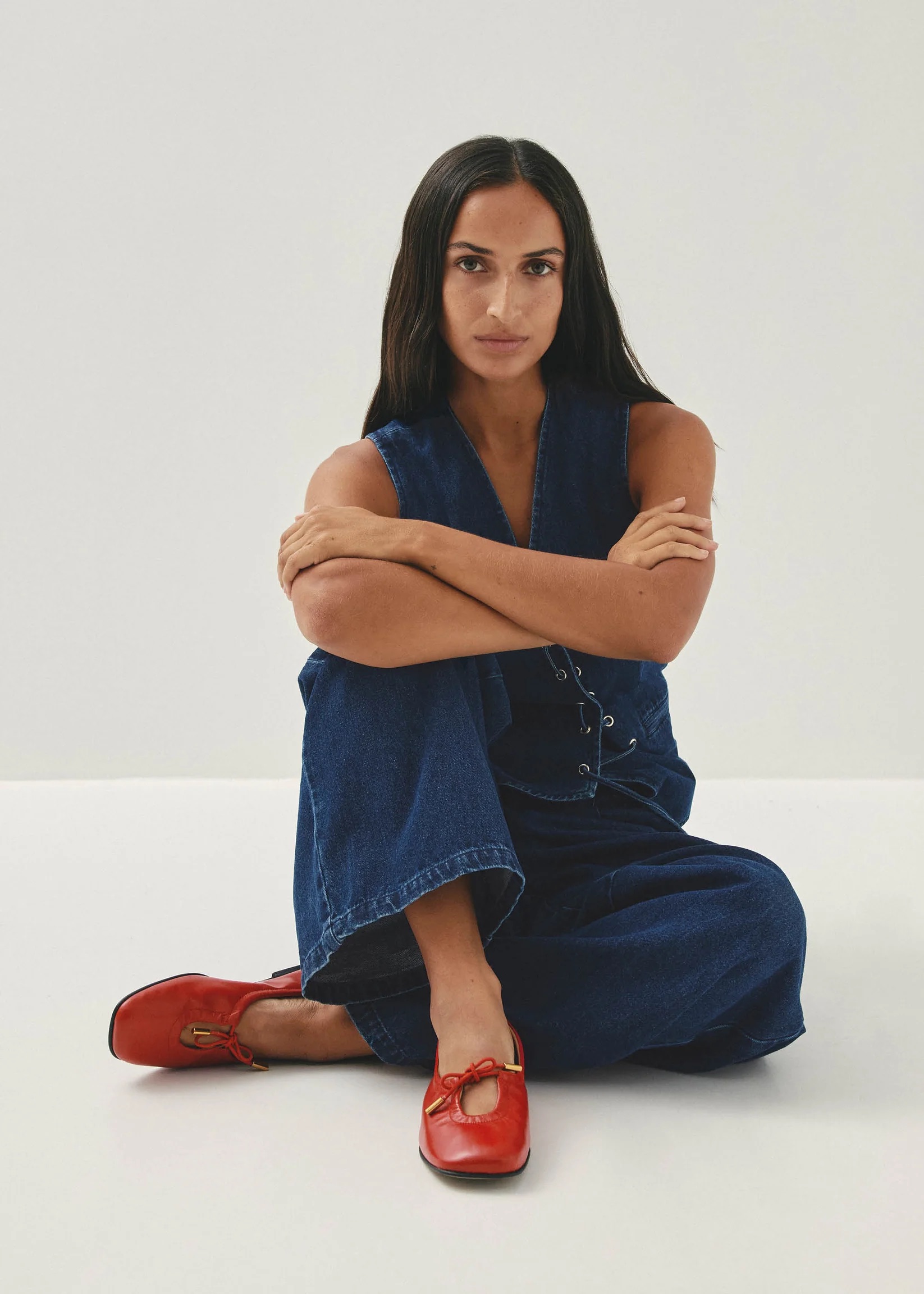 Person with long dark hair sits cross-legged on the floor, wearing a blue sleeveless jumpsuit and red shoes, against a plain white background.
