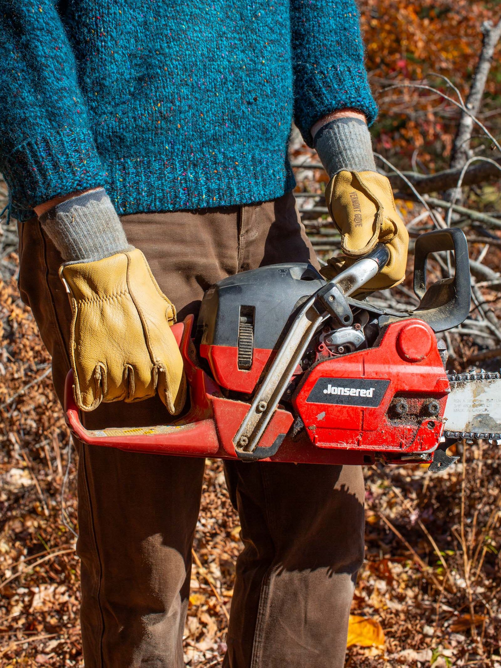 Person in a blue sweater and brown pants holding a red chainsaw, wearing yellow gloves, standing in a forest with autumn leaves.