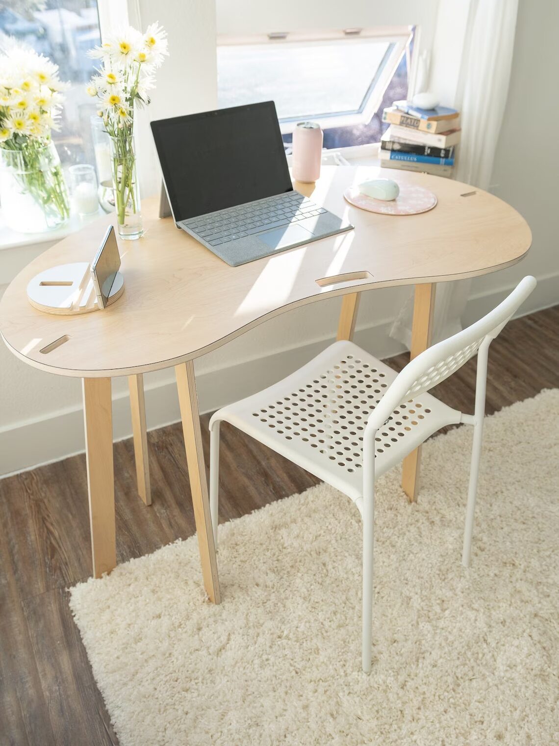 A light wood desk with a white chair, laptop, tablet, flowers in vases, a drink, books, and a plate on a white rug in a sunlit room.