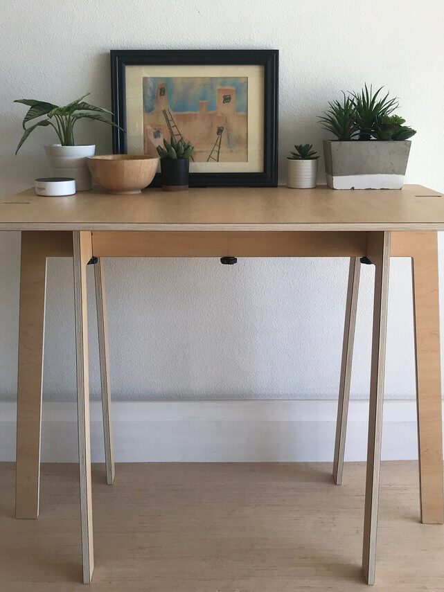 A small wooden table with potted plants, a wooden bowl, and a framed artwork arranged against a plain white wall.