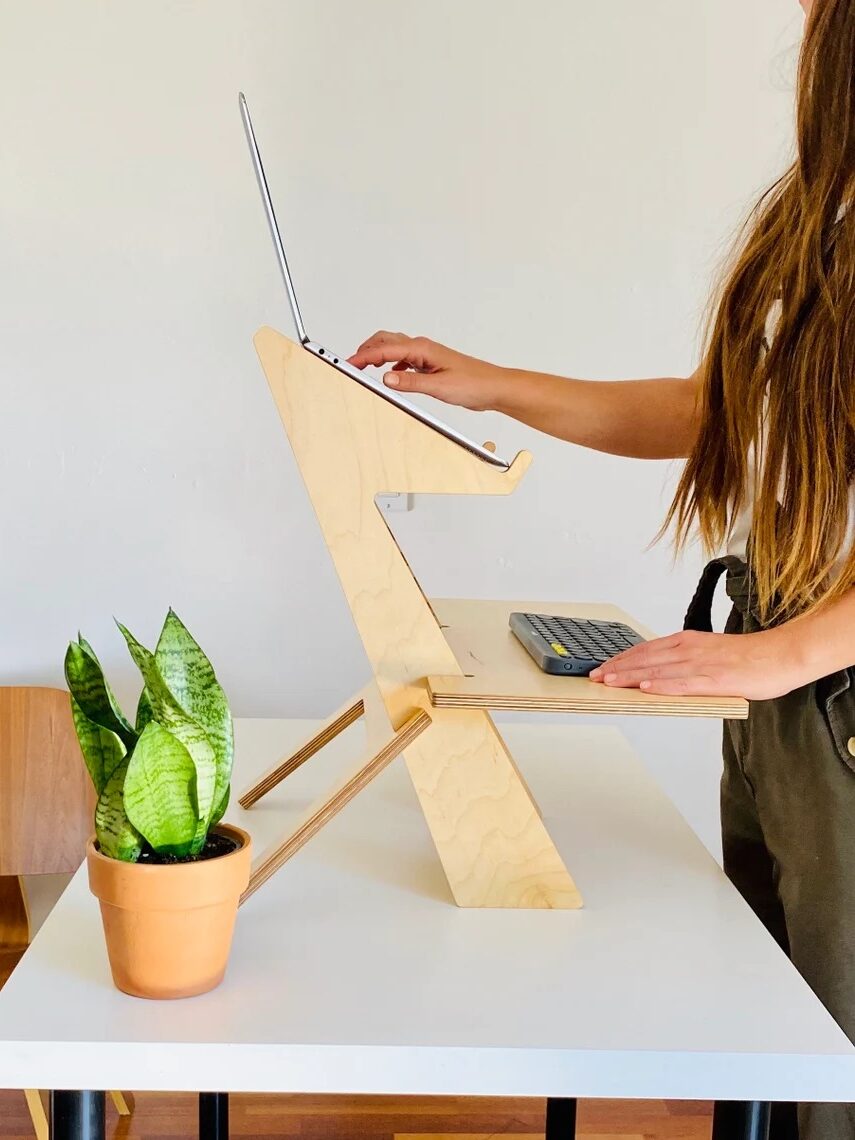 A person uses a wooden standing desk converter on a white table, with a laptop on top and a potted plant and wireless keyboard nearby. A wooden chair is beside the table.