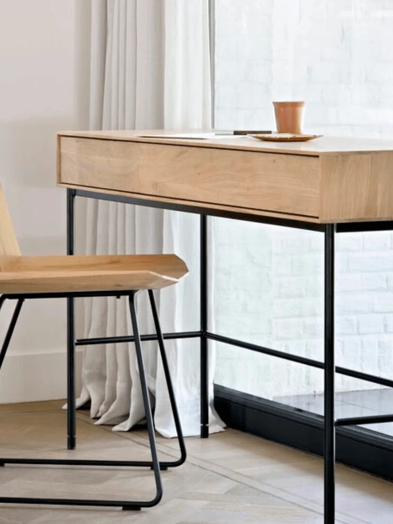A minimalist wooden desk and chair with black metal legs are positioned by a window with sheer white curtains. A notebook and a cup sit on the desk.