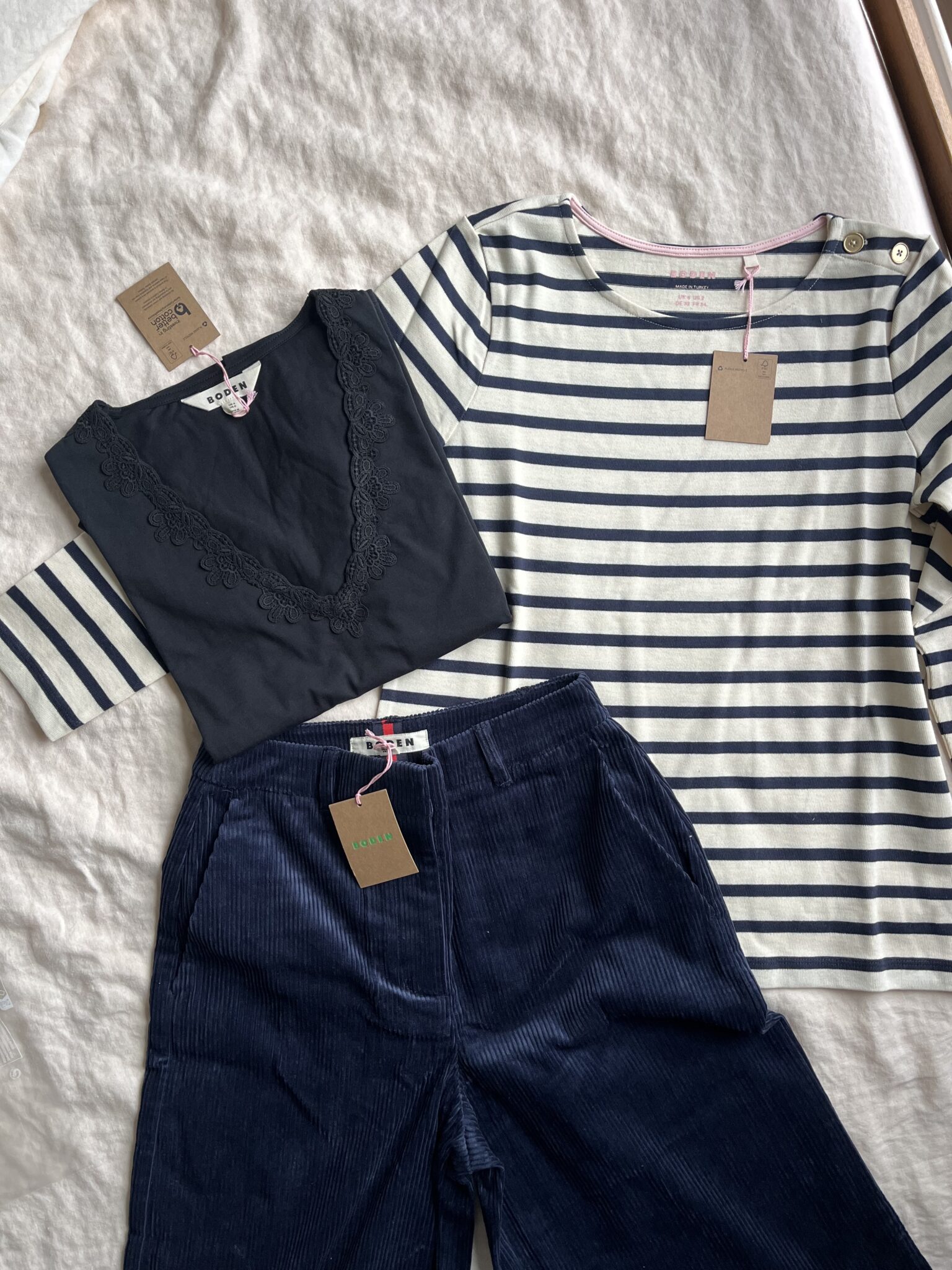 Three items of new clothing with tags: a navy V-neck top with embroidery, a white and navy striped long-sleeve shirt, and navy corduroy pants, arranged on a beige fabric background.