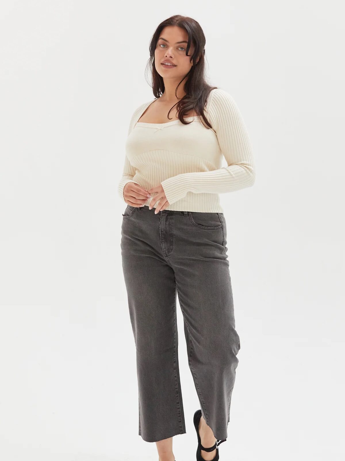 A woman stands against a plain white background wearing a cream-colored long-sleeve top, gray jeans, and black flats.