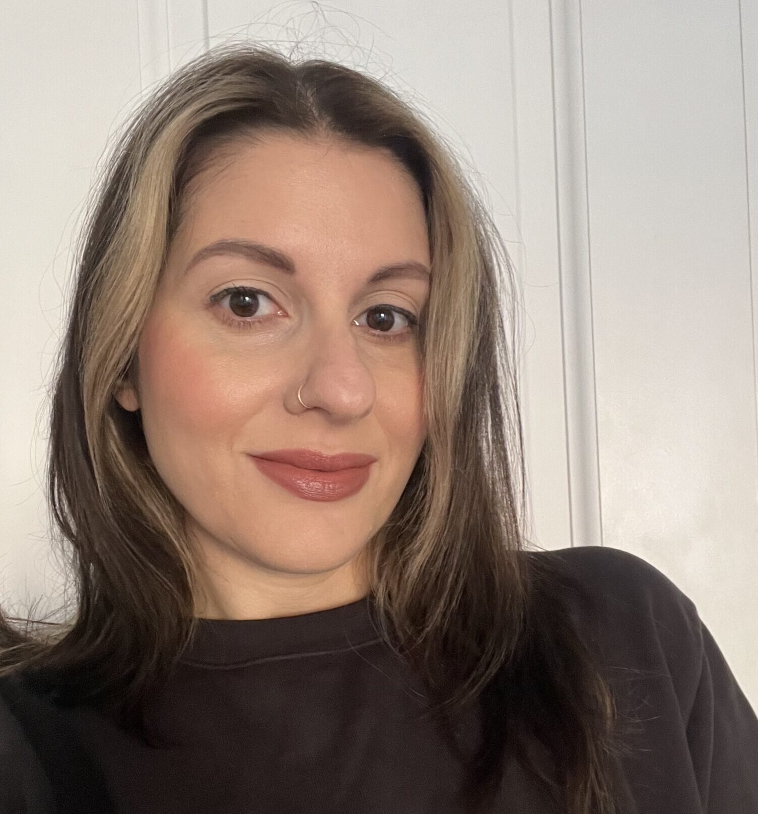 A woman with straight, shoulder-length brown hair with blonde highlights, wearing a black top, poses in front of a white paneled wall.