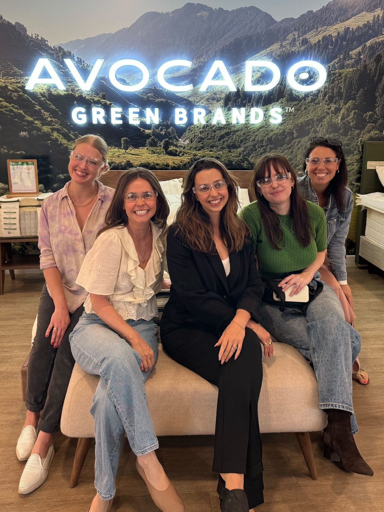 Five members of The Good Trade team wearing safety glasses pose on a bench in front of an "Avocado Green Brands" sign at the brand's factory in Fullerton California.