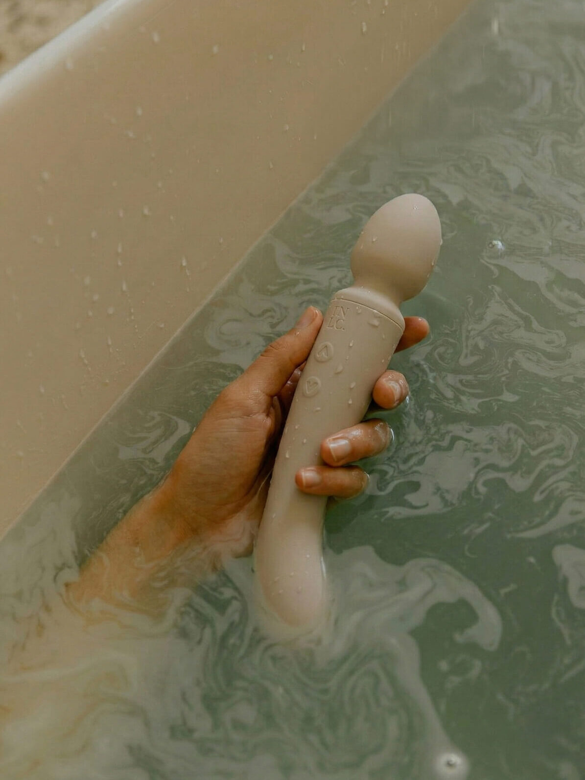 A hand holding a waterproof personal massager partially submerged in bathwater.