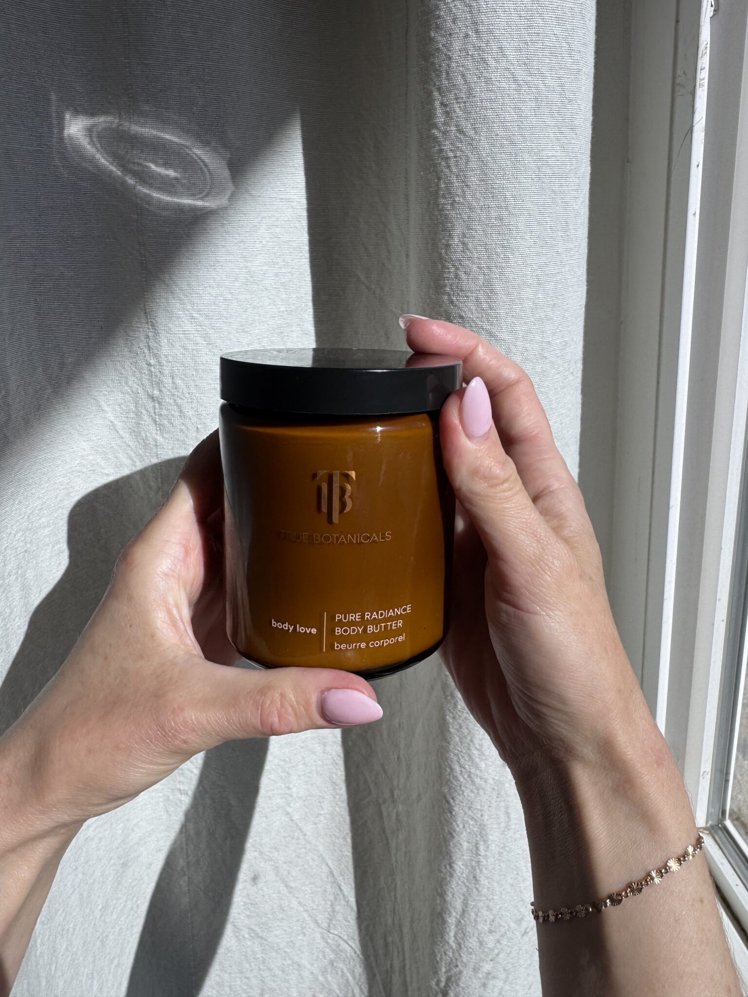 Person holding a jar of True Botanicals Pure Radiance Body Butter against a light-colored curtain with sunlight casting shadows.