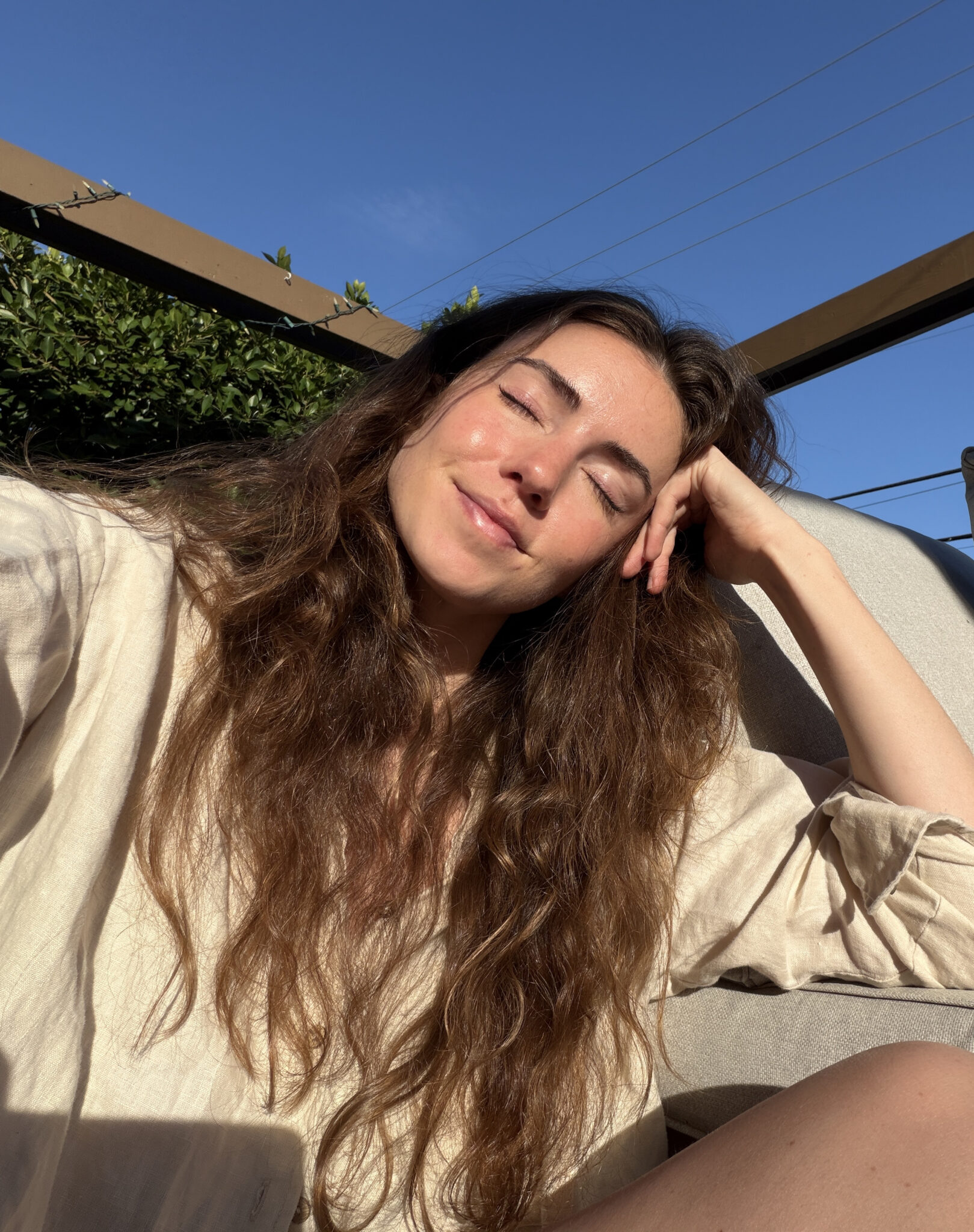 A woman with long brown hair sits outdoors on a sunny day, eyes closed and smiling, leaning her head on her hand.
