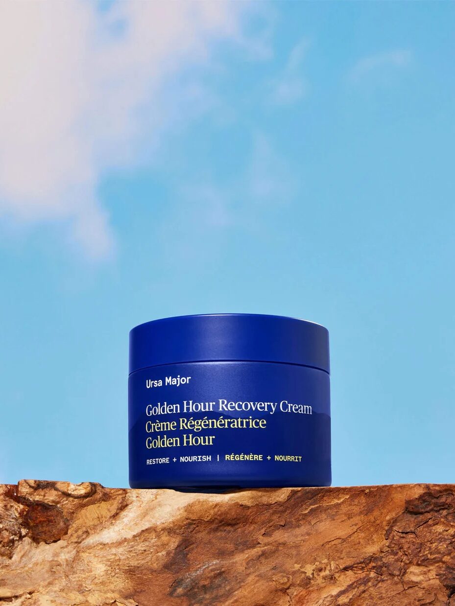 A blue jar of Ursa Major Golden Hour Recovery Cream sits on a rock ledge against a blue sky with clouds.