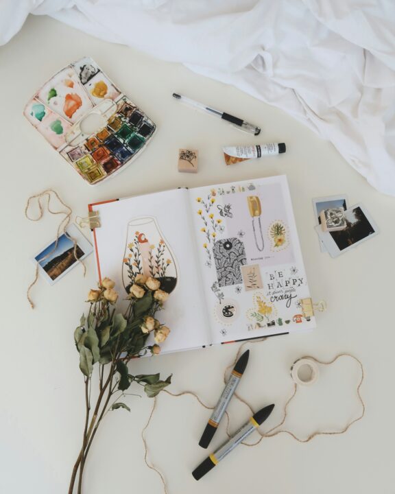An open sketchbook with drawings and stickers lies on a desk surrounded by markers, dried flowers, watercolor paints, photos, string, and art supplies.