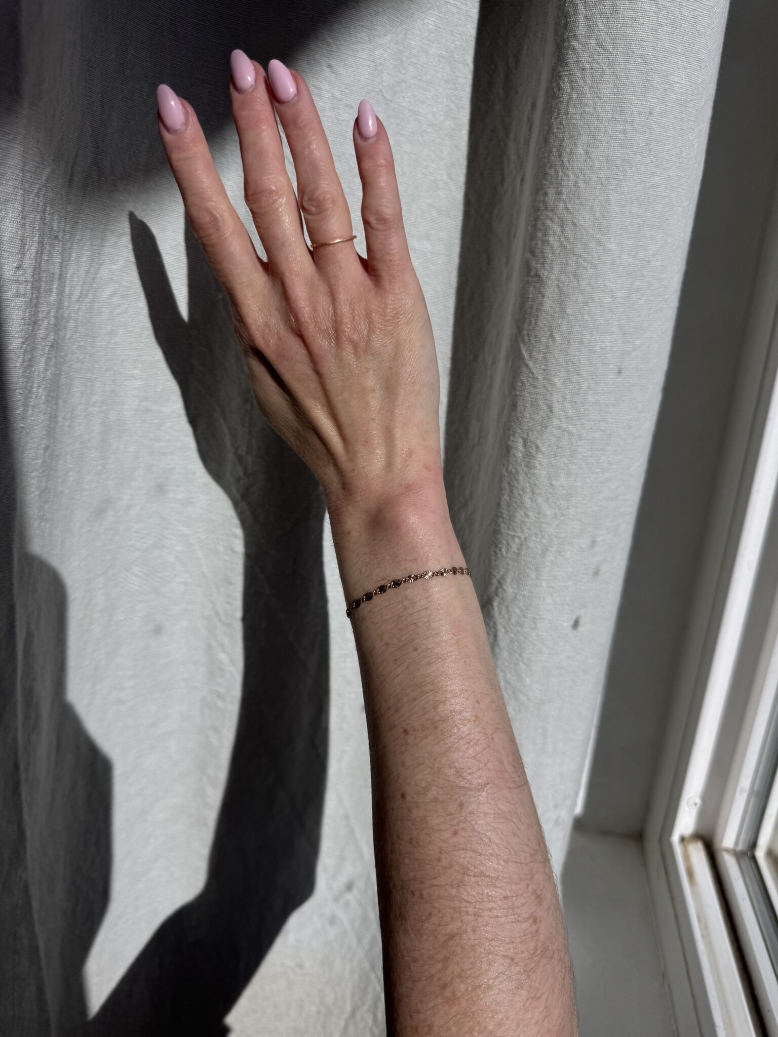 A hand with light pink nail polish and a thin bracelet is raised against a sunlit white curtain, casting a shadow.