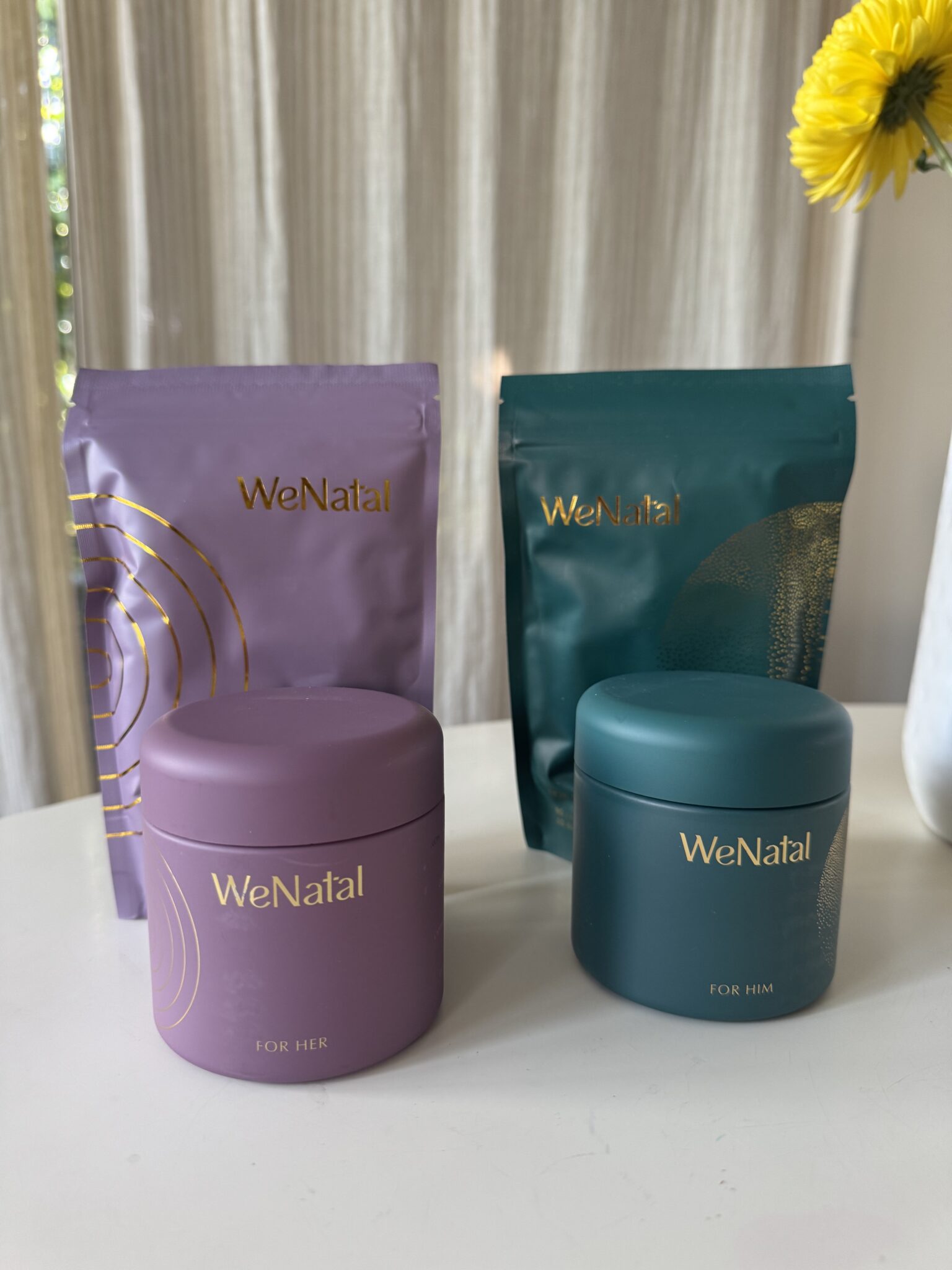 Two WeNatal supplement containers and two refill pouches are displayed on a white surface with a flower vase in the background.