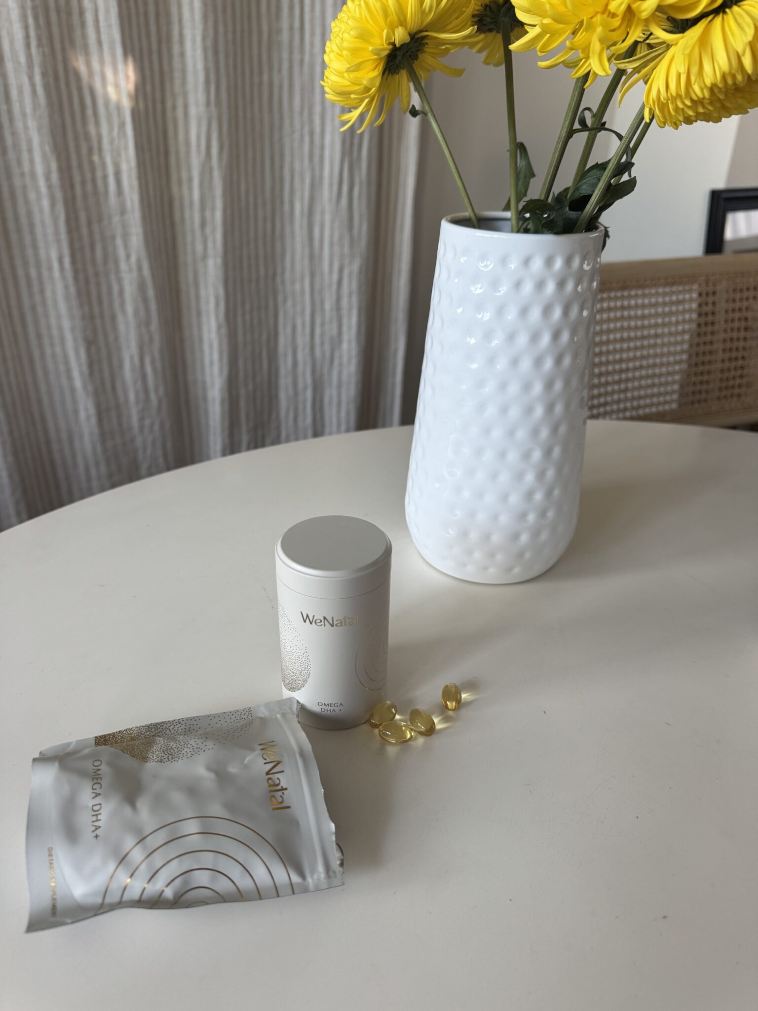 A white vase with yellow flowers sits on a round table next to a supplement container, an opened packet, and several yellow capsules.