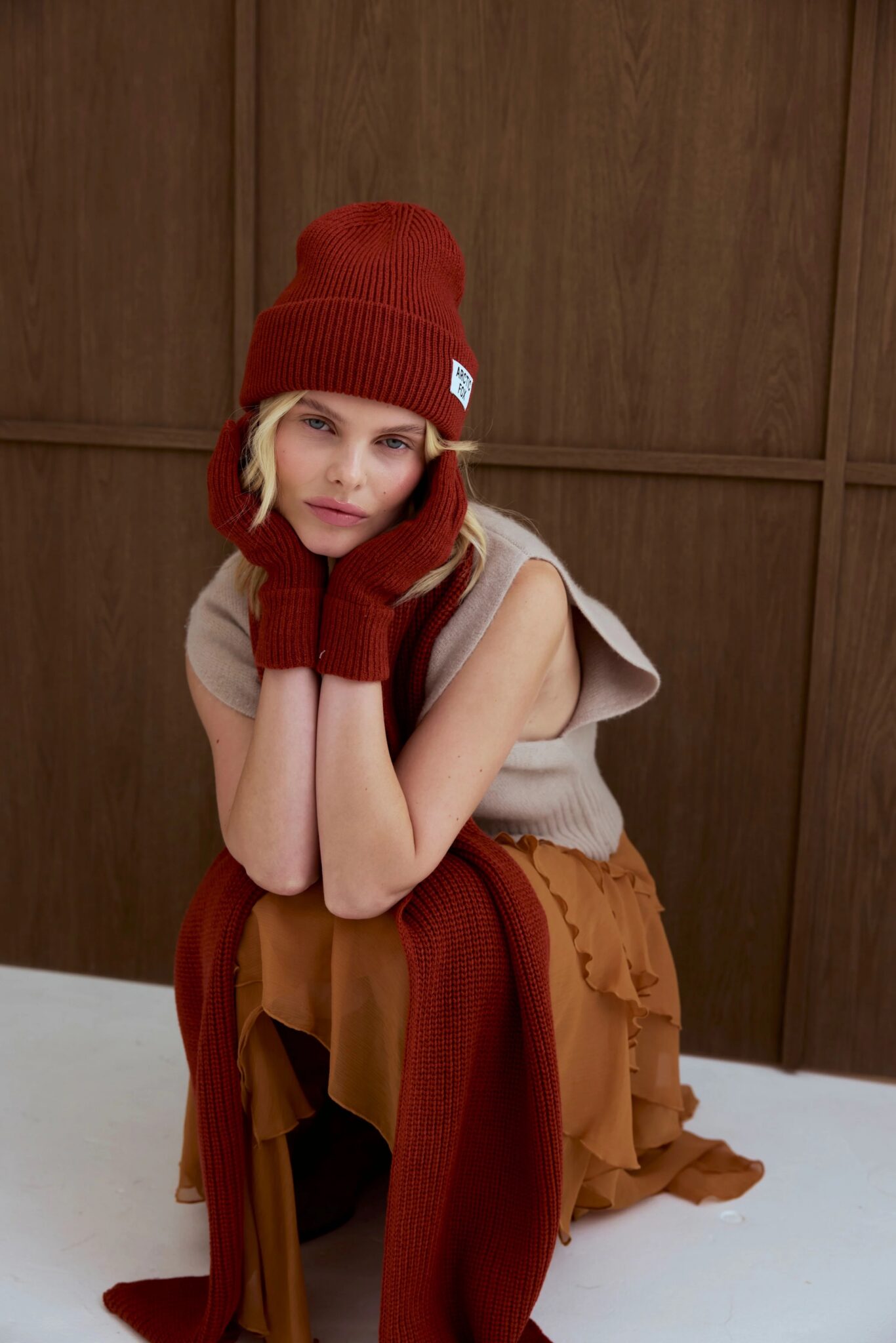 A person wearing a beige sleeveless top, ruffled brown skirt, and matching red knit hat, gloves, and scarf sits indoors with their chin resting on their hands.