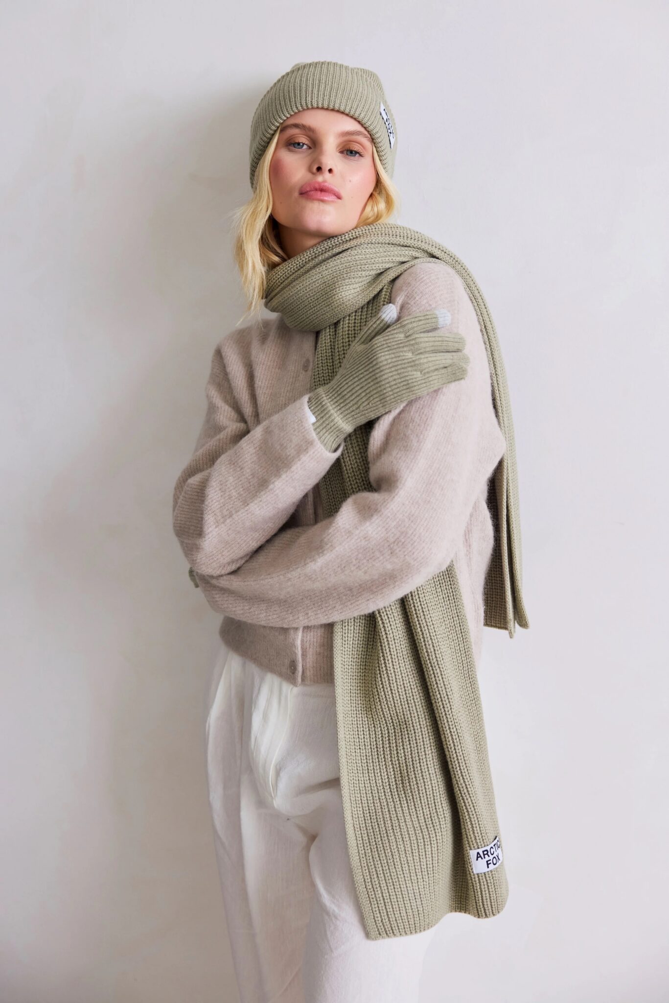 A woman wearing a beige knit hat, matching scarf, and gloves, with a light beige sweater and white pants, stands against a plain light background.