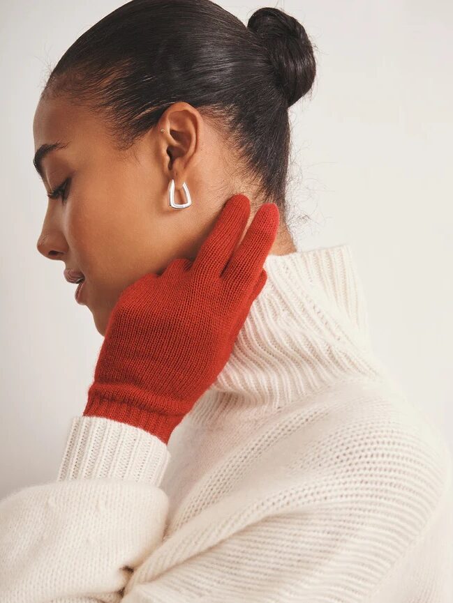 A woman in a white turtleneck sweater wears red gloves and silver geometric earrings, facing sideways with her gloved hand touching her cheek.