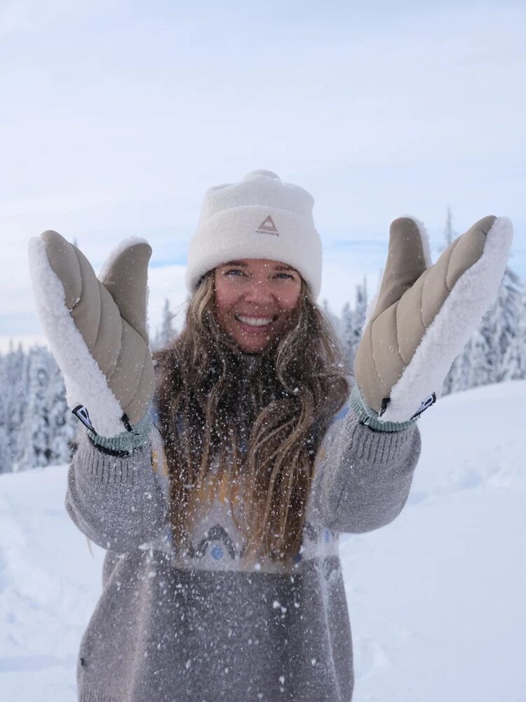 A woman wearing a winter hat, sweater, and mittens stands outdoors in a snowy landscape, smiling and holding her gloved hands toward the camera as snow falls.