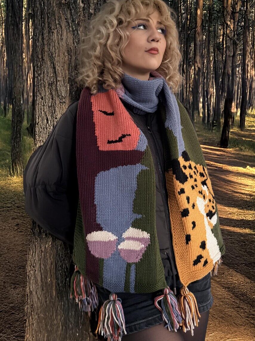 A person with curly blonde hair wears a colorful patterned scarf and stands in a sunlit forest, leaning against a tree.