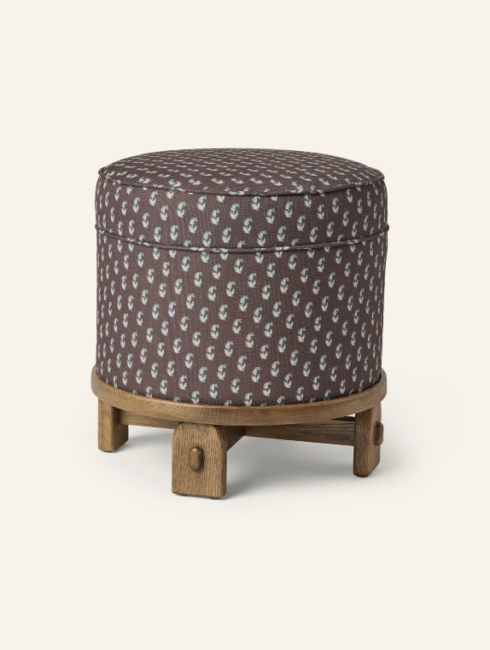Round, cushioned ottoman with a patterned fabric cover in brown and blue, set on a light wooden base with four short legs.