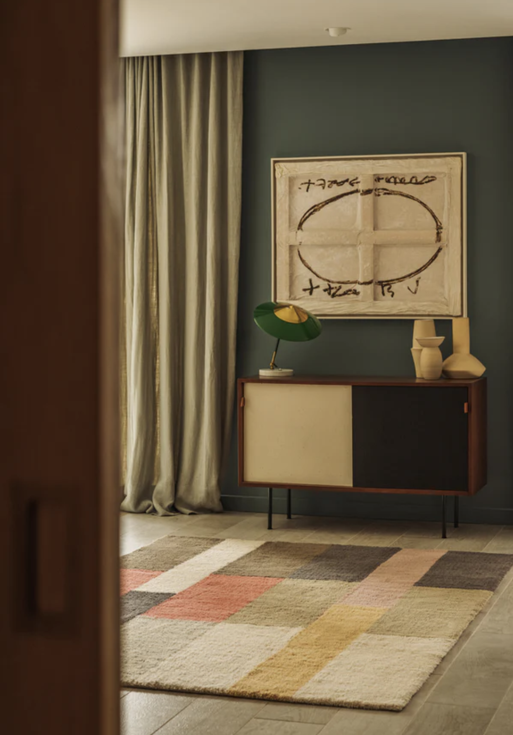Interior with a mid-century modern sideboard, abstract wall art, green lamp, two vases, neutral curtains, and a colorful geometric rug on a tiled floor.