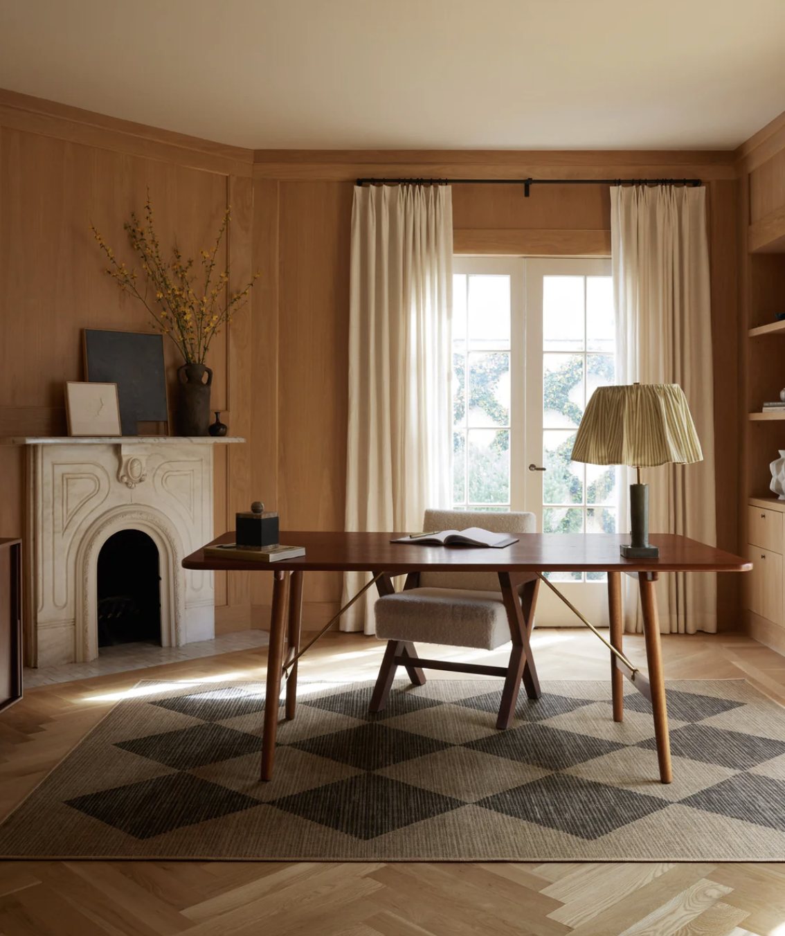 A wooden desk with a chair, table lamp, and books is centered on a patterned rug in a room with wooden walls, a fireplace, and a window with curtains.