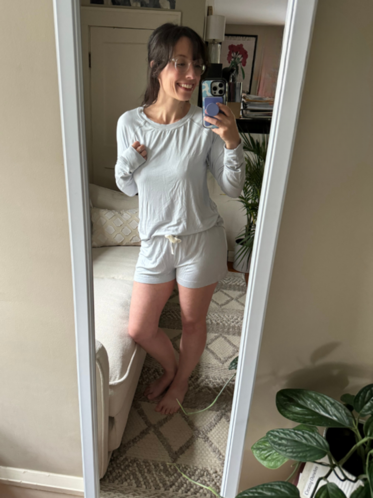 Woman taking a mirror selfie in a light-colored long sleeve top and shorts, standing barefoot in a cozy, well-lit living room with plants and neutral decor.