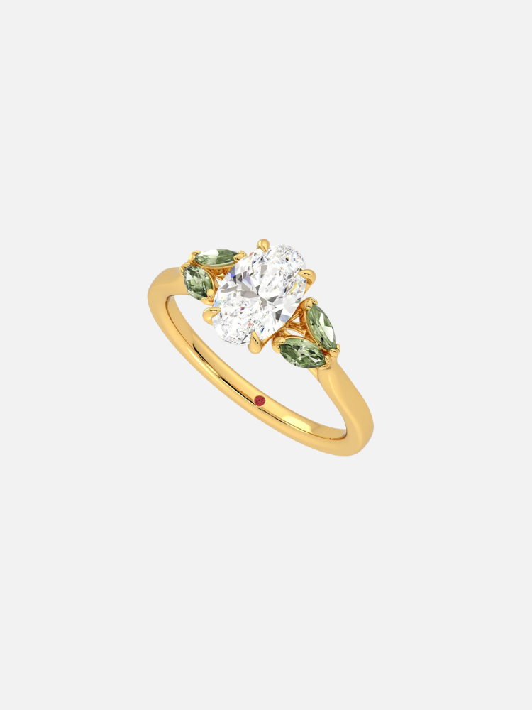 Gold ring with three stones: a central oval white gem and two green gems on either side, set on a gold band with a small red dot detail.