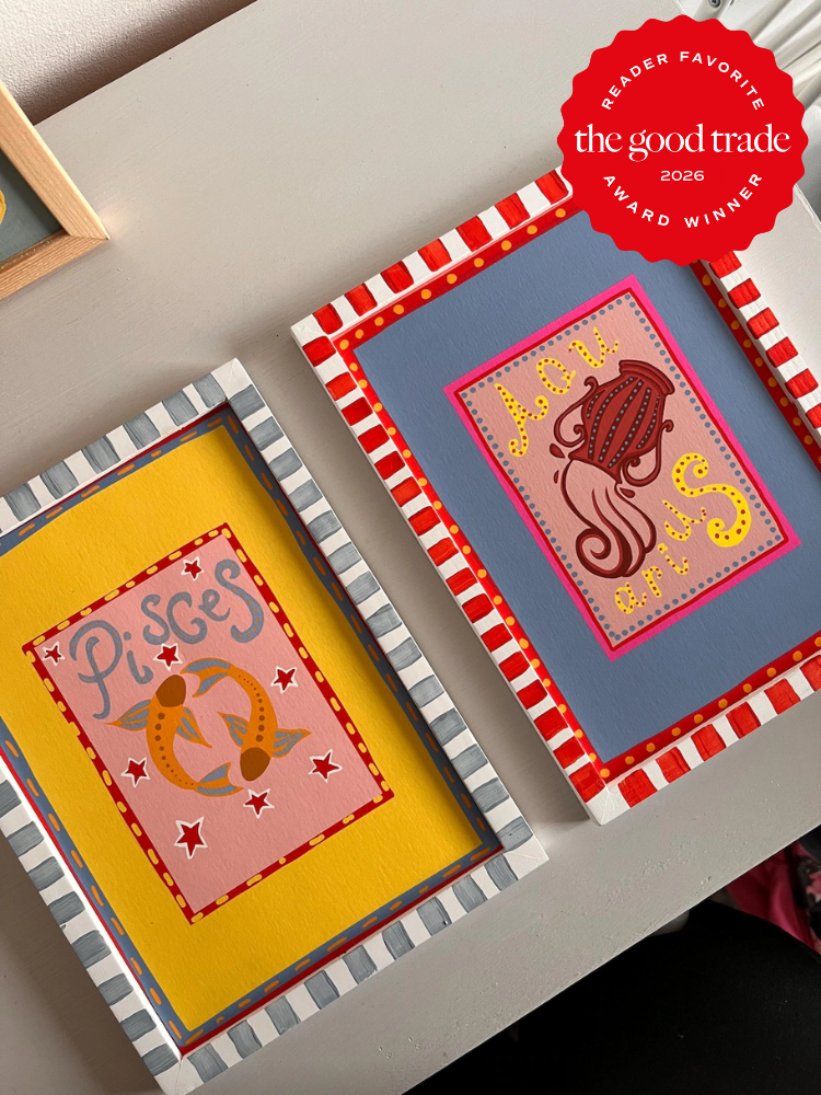 Two colorful, framed zodiac paintings—Pisces on yellow and Aquarius on blue—sit on a white desk. A "The Good Trade Award Winner 2026" badge is in the top right corner.