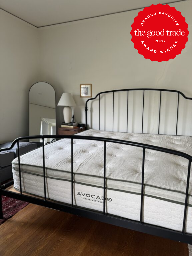 A bedroom with a black metal bed frame, an Avocado organic mattress, a nightstand with a lamp, a mirror, and a red "The Good Trade 2026 Award Winner" badge in the upper right corner.