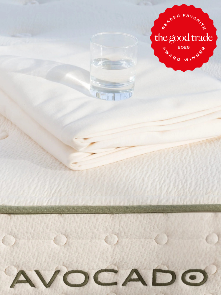 A glass of water sits on folded white bedding atop an Avocado mattress. A red badge in the corner reads, "The Good Trade Reader Favorite Award Winner 2026.
