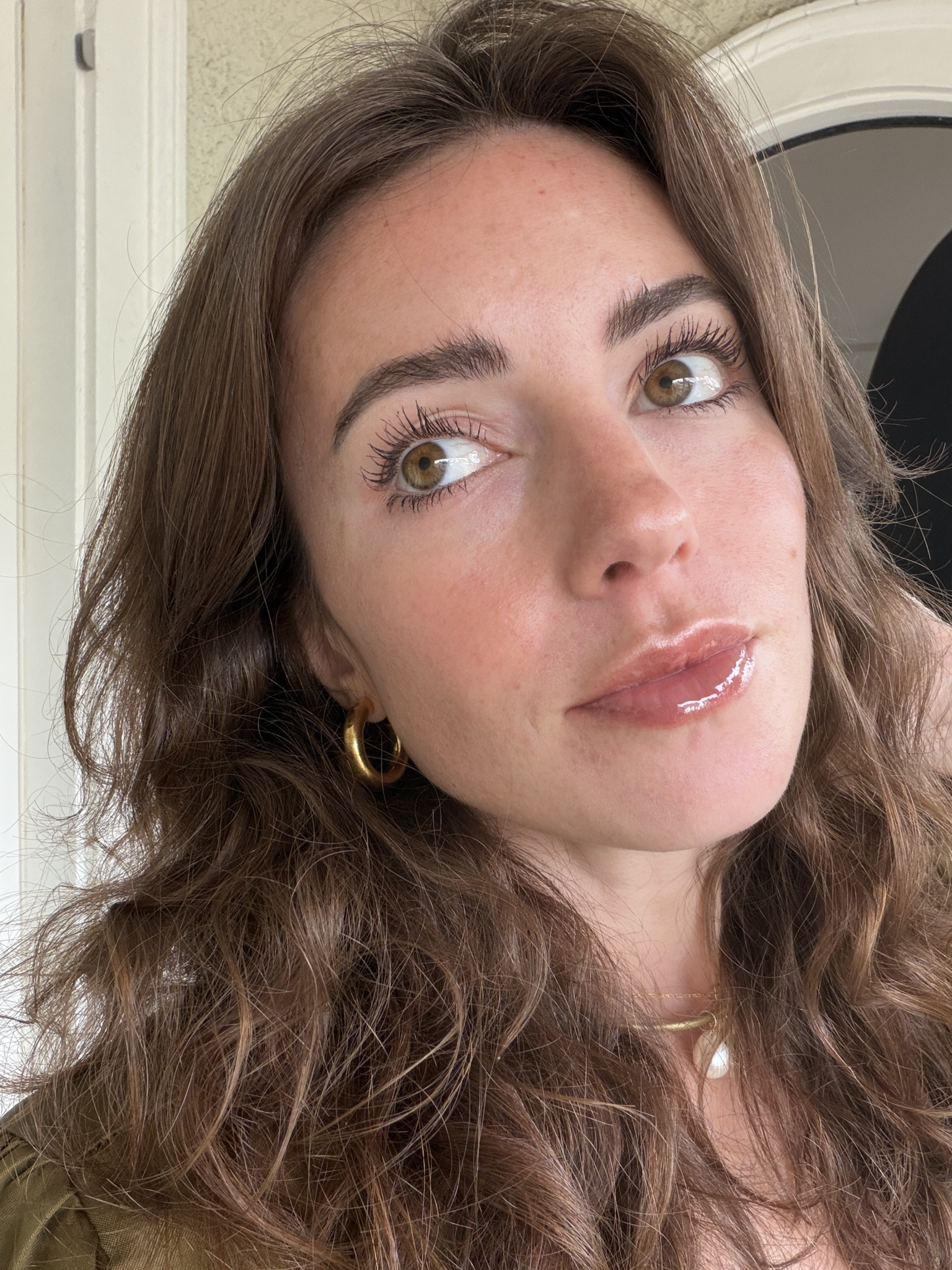 A woman with wavy brown hair, glossy lips, and gold hoop earrings looks slightly upward indoors, with natural light illuminating her face.