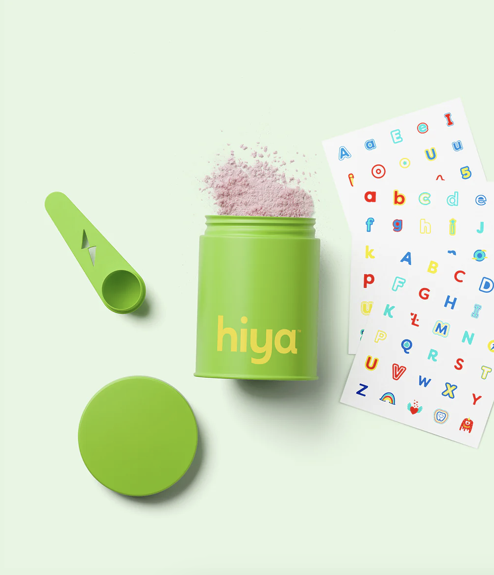 A green container labeled "hiya" with pink powder inside, a matching scoop, a detached lid, and a colorful alphabet sticker sheet on a light green background.