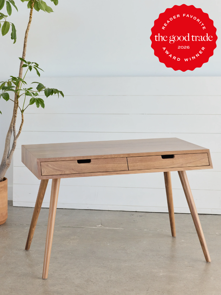 A wooden desk with two drawers stands on angled legs indoors; a potted plant is nearby and a red award badge is displayed in the top right corner.