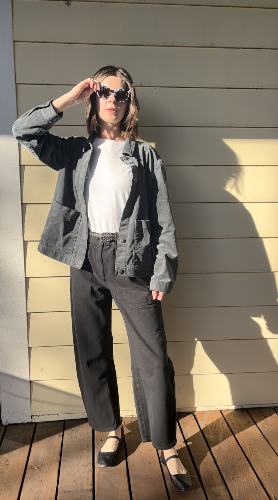 Person wearing sunglasses, a gray jacket, white t-shirt, black pants, and black shoes stands on a wooden deck in sunlight, casting a shadow on a light-colored wall.