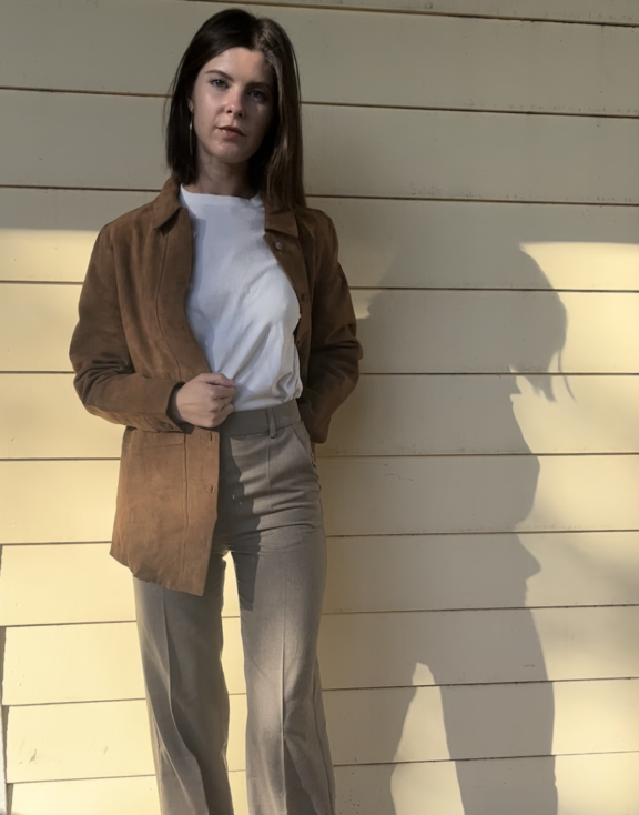 A person stands on a wooden deck against a cream paneled wall, wearing a brown jacket, white t-shirt, beige pants, and black shoes in sunlight.